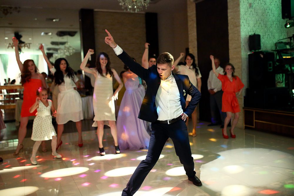 A groom dances with a disco pose in front of a group of female wedding guests