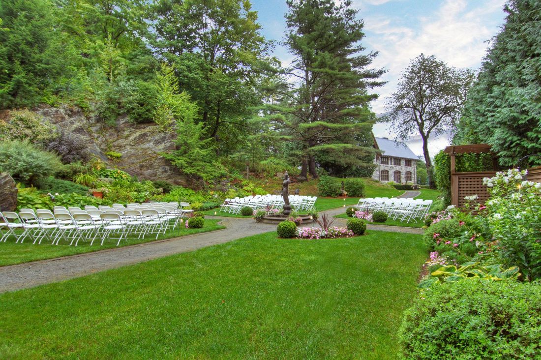 White chairs are spread out on a lawn, on either side of a dirt path leading to a stone building.