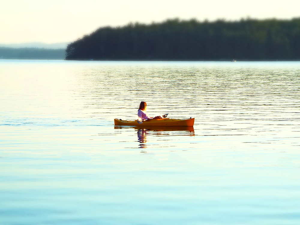 A person paddles a yellow kayak on a calm lake in New Hampshire