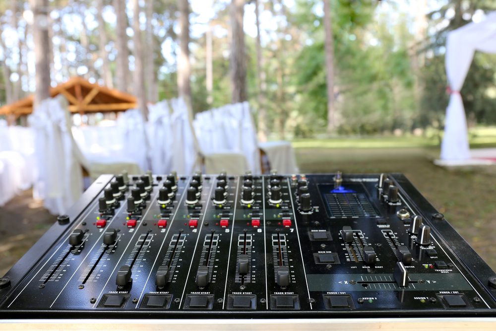 A dj mixer set up at an outdoor wedding, with white fabric draping chairs and tall trees in the background.