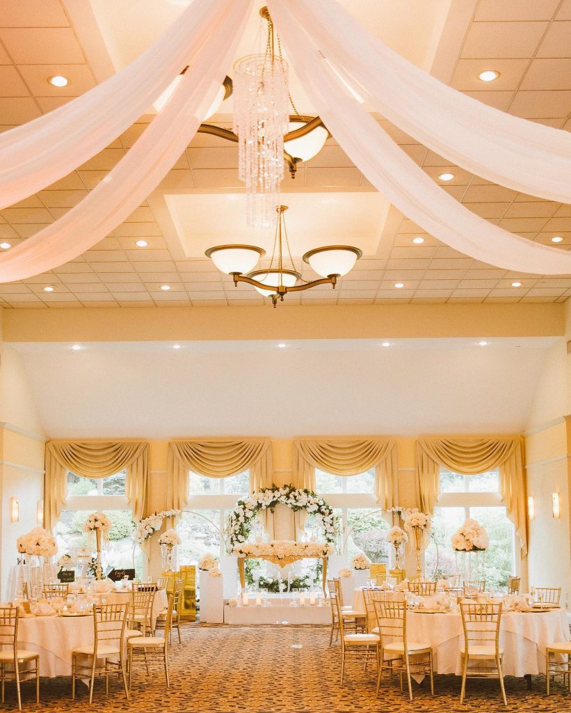 A ballroom glows with warm light, with white fabric and chandeliers hanging from a high ceiling at a wedding venue in NH.